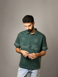 A man wearing a dark green polo shirt with 'Quiet Wins' printed in white script across the chest, paired with light blue jeans, standing against a neutral gray studio background.
