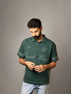 A man wearing a dark green polo shirt with 'Quiet Wins' printed in white script across the chest, paired with light blue jeans, standing against a neutral gray studio background.
