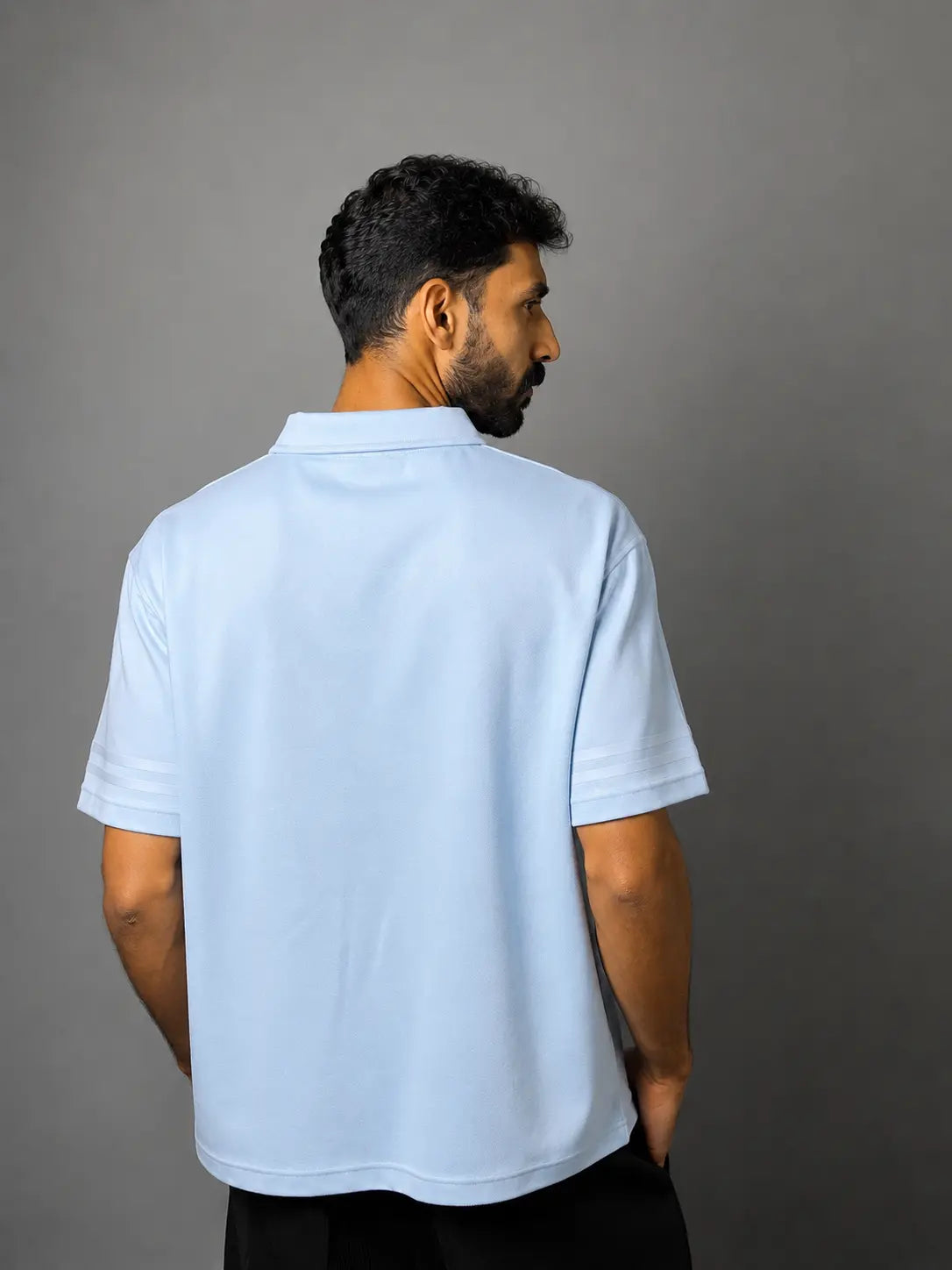 A man wearing a light blue short-sleeve polo shirt, shown from the back against a solid dark gray studio background.