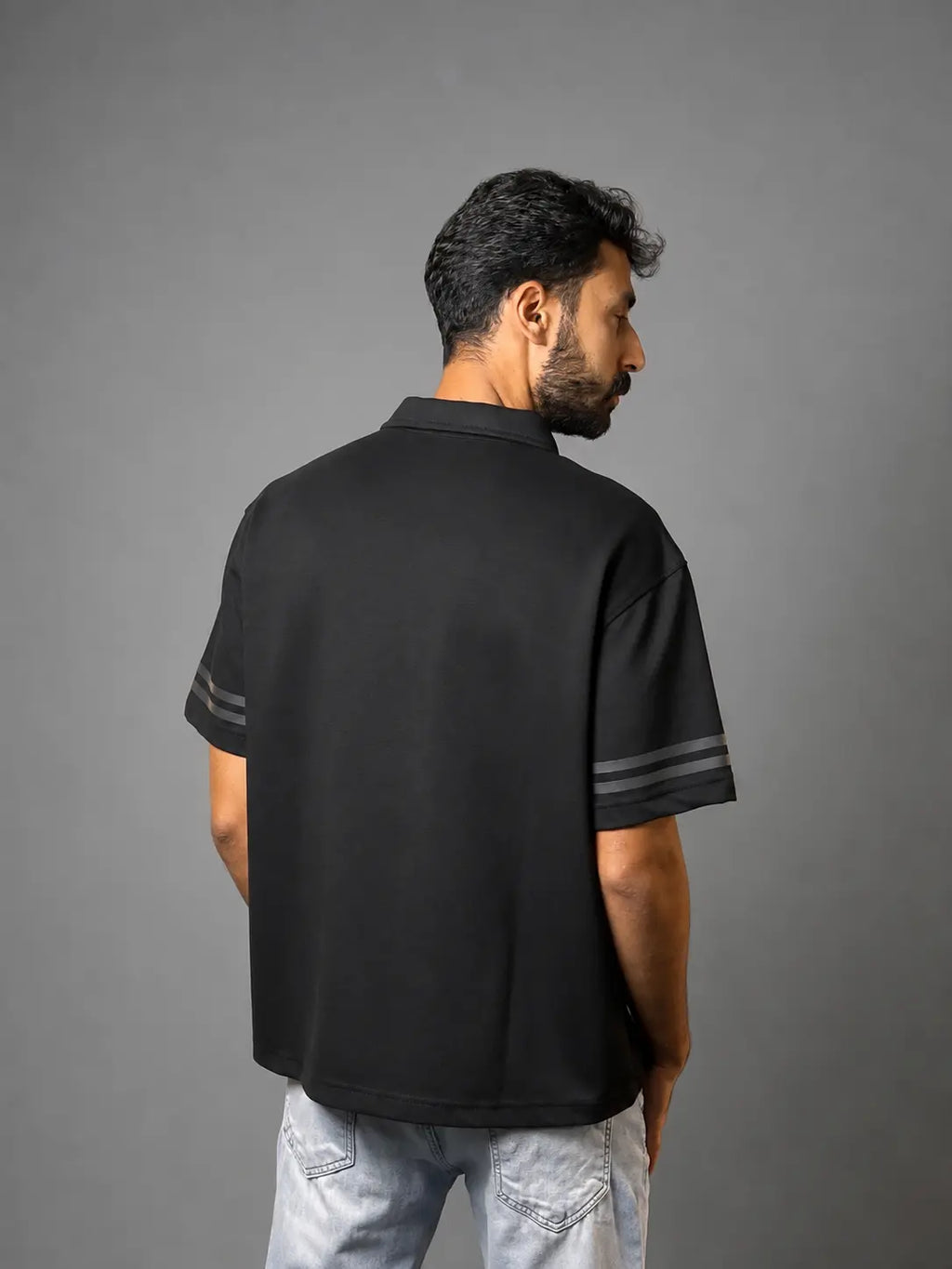 A man shown from the back wearing a black short-sleeve polo shirt with gray stripes on the sleeve cuffs, paired with light wash jeans, against a solid gray studio background.