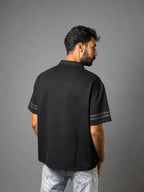 A man shown from the back wearing a black short-sleeve polo shirt with gray stripes on the sleeve cuffs, paired with light wash jeans, against a solid gray studio background.