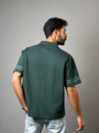 A man wearing a dark green short-sleeve polo shirt with light gray sleeve stripes, shown from the back against a solid gray background.