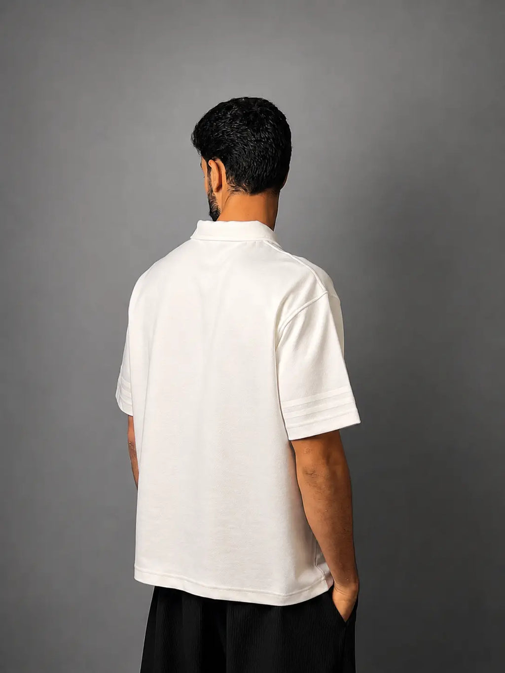 A man viewed from behind wearing a white short-sleeve polo shirt and black pants, standing against a solid gray studio background.