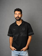 A man wearing a black short-sleeve polo shirt with gray sleeve stripes and gray script lettering on the front, paired with light blue jeans, standing against a solid gray background.