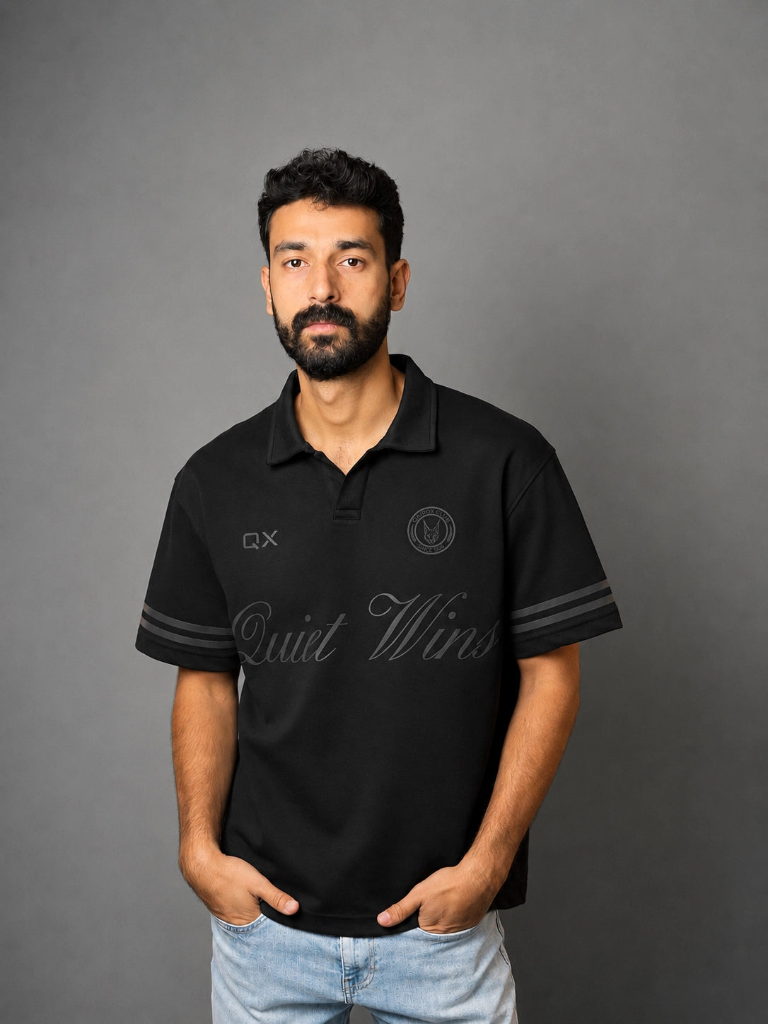 A man wearing a black short-sleeve polo shirt with gray sleeve stripes and gray script lettering on the front, paired with light blue jeans, standing against a solid gray background.