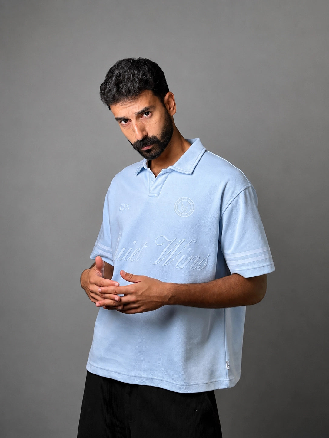 A man wearing a light blue polo shirt with white script embroidery across the chest, posed against a solid gray studio background.