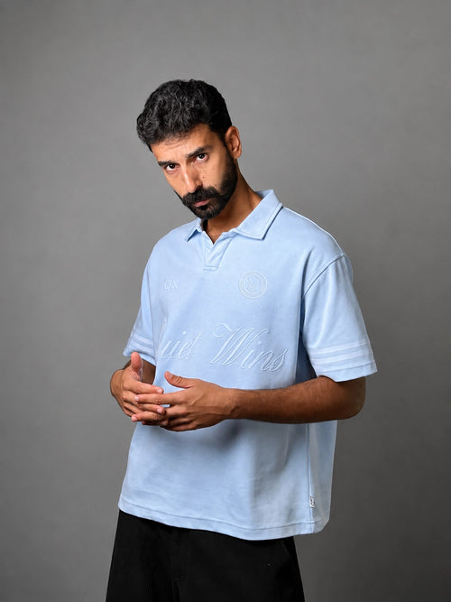 A man wearing a light blue polo shirt with white script embroidery across the chest, posed against a solid gray studio background.