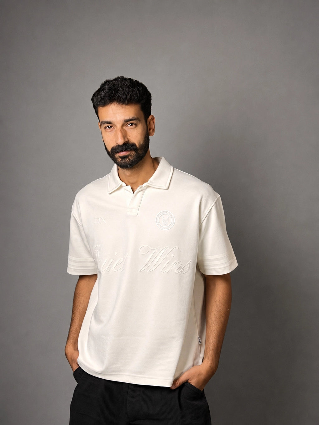 A man with a beard modeling a white short-sleeve polo shirt with tonal embroidery, standing against a solid gray background.