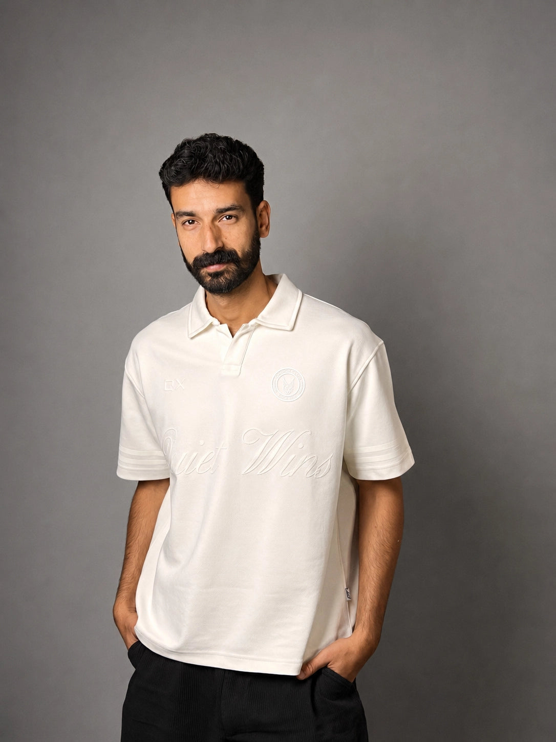 A man with a beard modeling a white short-sleeve polo shirt with tonal embroidery, standing against a solid gray background.