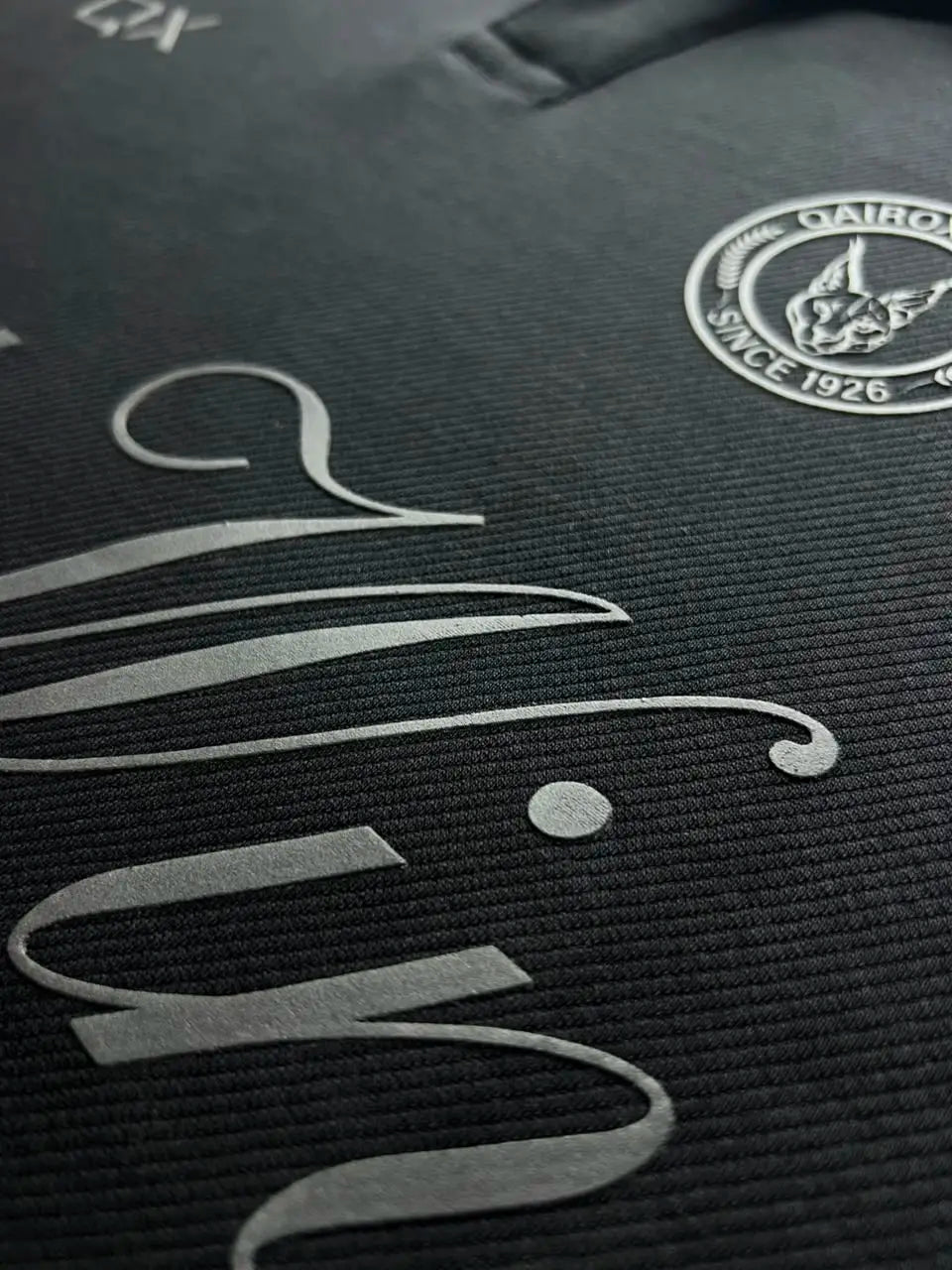 Close-up of a black textured garment with a silver-toned script logo and a circular Cairo FC crest on the chest.