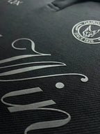 Close-up of a black textured garment with a silver-toned script logo and a circular Cairo FC crest on the chest.