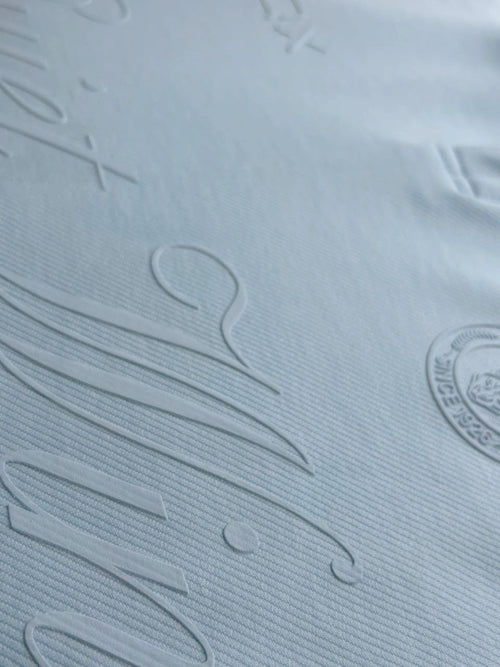 Close-up of a light blue textured fabric with embossed script lettering and a circular logo on the right side.