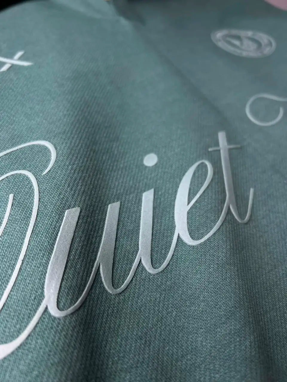 Close-up of a dark teal-green fabric with a large white cursive script reading "Quiet" across the front.