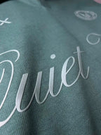 Close-up of a dark teal-green fabric with a large white cursive script reading "Quiet" across the front.